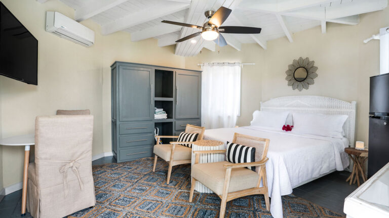 King bed with ceiling fan, accent chairs, and wardrobe in the Parrot Fish Suite at Arawak Beach Club in Anguilla